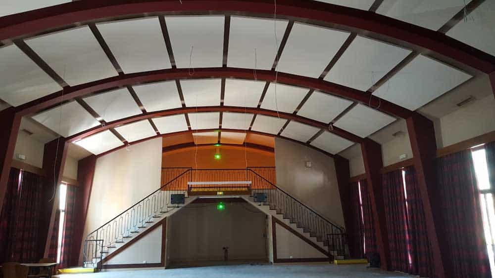 Salle de Réception avec Escalier Double et Plafond Voûté Salle polyvalente avec escalier double, plafond voûté à poutres bordeaux et panneaux acoustiques blancs.