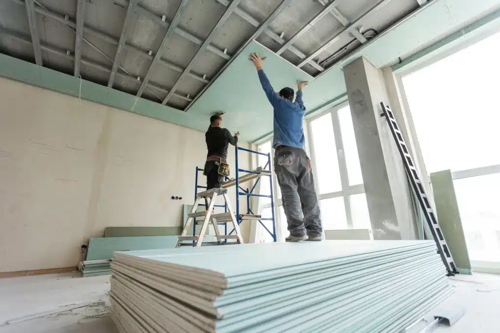 Installation de Plafond en Placo : Artisans en Chantier Artisans installant des plaques de plâtre hydrofuges au plafond d'une pièce en rénovation avec échafaudage et escabeau.